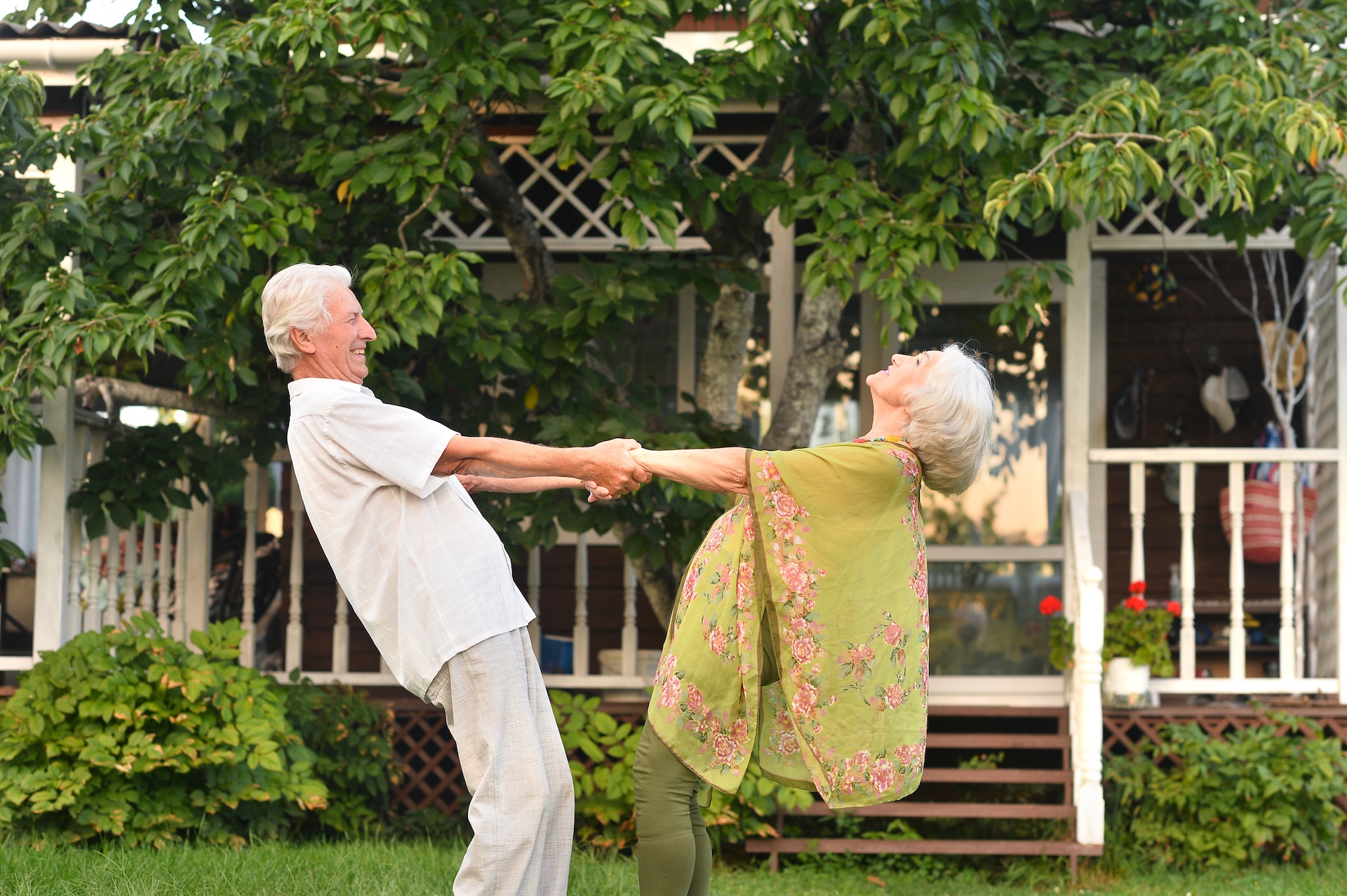 Ein älteres Paar tanzt lachend vor einem Haus im Garten.